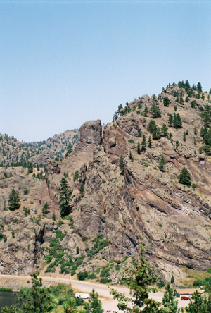 [It appears part of the stone near the top edge of a large rock outcropping has split. While this entire rock scene is mostly brown, there are a few evergreens growing on it. There is a roadway at the bottom of the rock.]